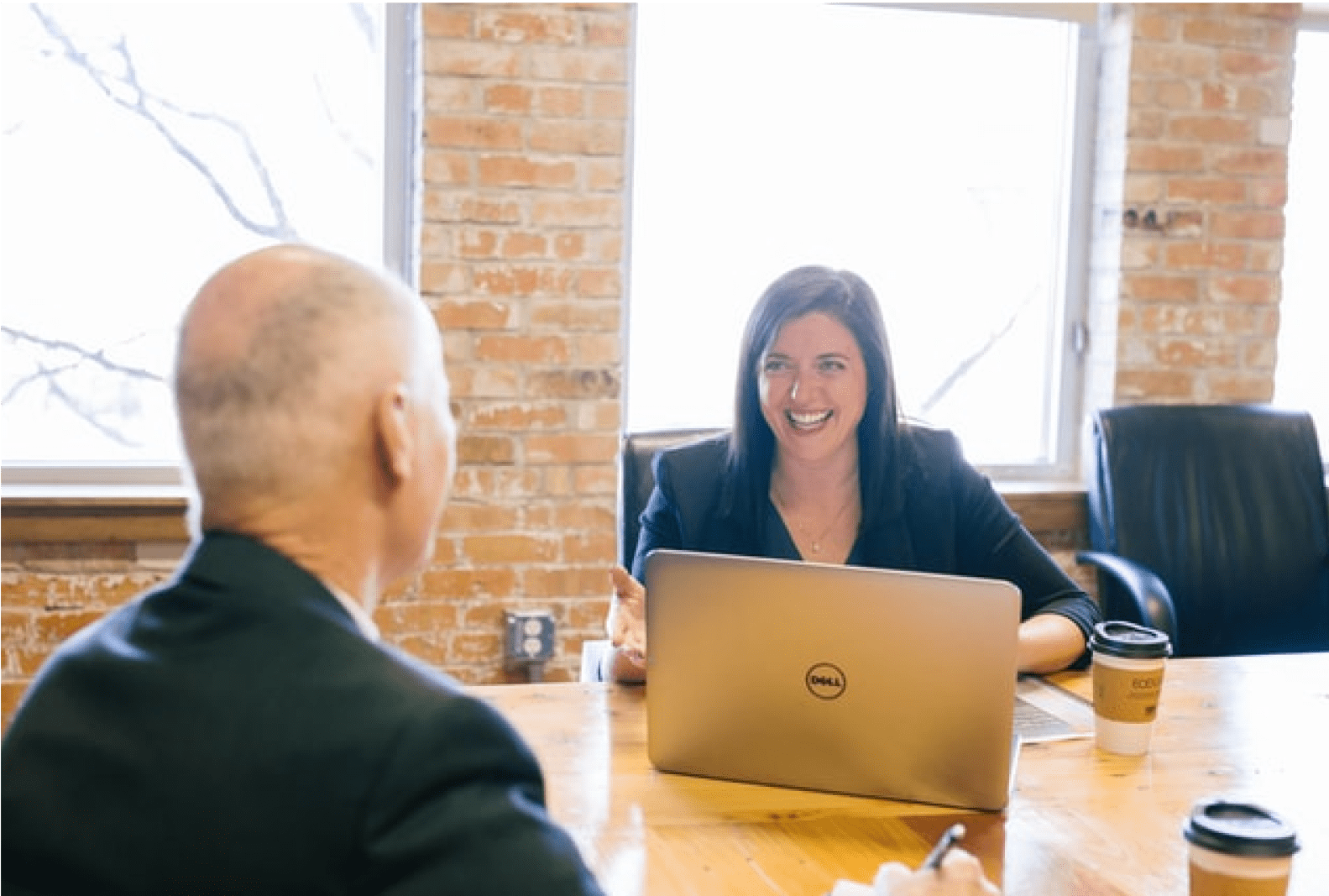 A smiling lady speaking in business attire with an elderly gentleman inside a brick meeting room.