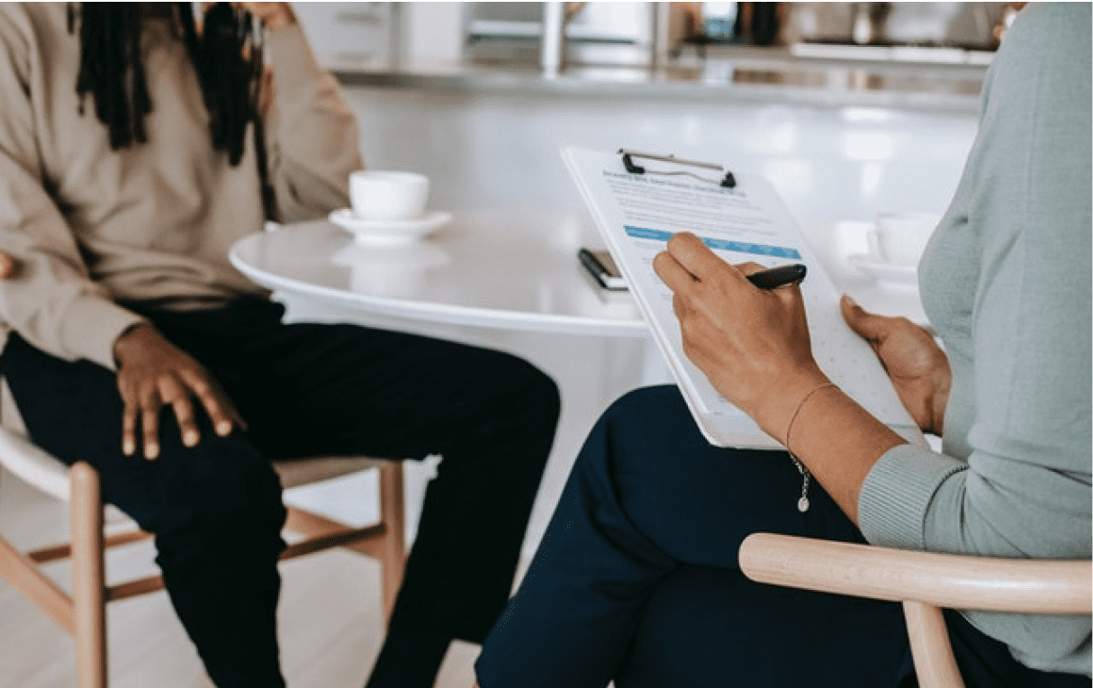 Two people having a discussion over coffee. One individual is holding a clipboard writing something down.